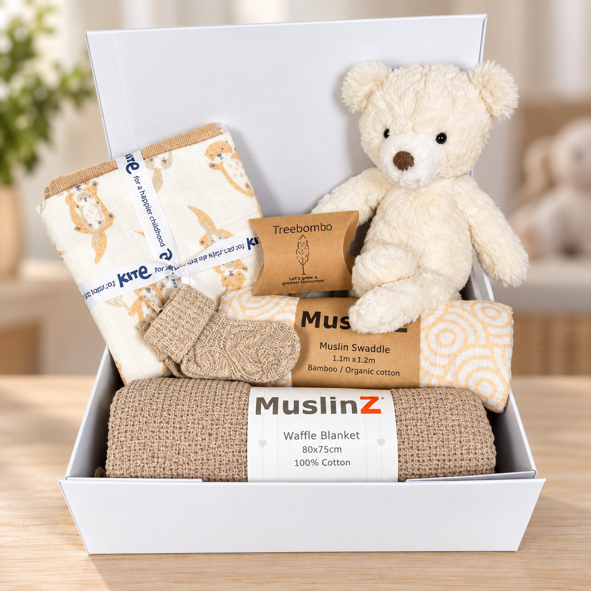 A baby gift set in a white box baby keepsake box featuring a MuslinZ waffle blanket in taupe, 3 organic cotton muslins, a muslin baby swaddle, a pair of baby socks, a Mary Meyer putty nursery teddy bear, and a Treebomb package, arranged on a wooden surface.
