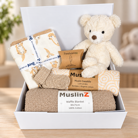A baby gift set in a white box baby keepsake box featuring a MuslinZ waffle blanket in taupe, 3 organic cotton muslins, a muslin baby swaddle, a pair of baby socks, a Mary Meyer putty nursery teddy bear, and a Treebomb package, arranged on a wooden surface.