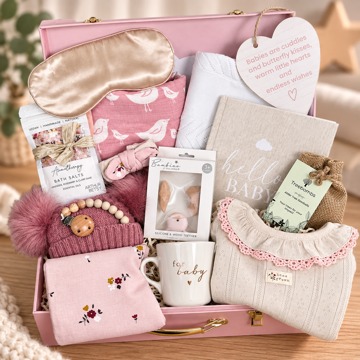 A pink baby gift hamper filled with baby accessories and treats, including a satin sleep mask, pink bird-print swaddle, a baby knit hat with pom-pom, a mug that says 'for baby', a heart-shaped nursery plaque, and packaged items, arranged on a wooden surface.