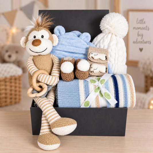 Baby Boy hamper containing a Wilberry knitted monkey baby soft toy, a blue baby dressing gown with a hood, a cotton striped baby blanket. A pair of crocheted baby booties with pompoms, a pair of cotton baby socks with elephant, a baby teething toy and a white baby pompom hat.