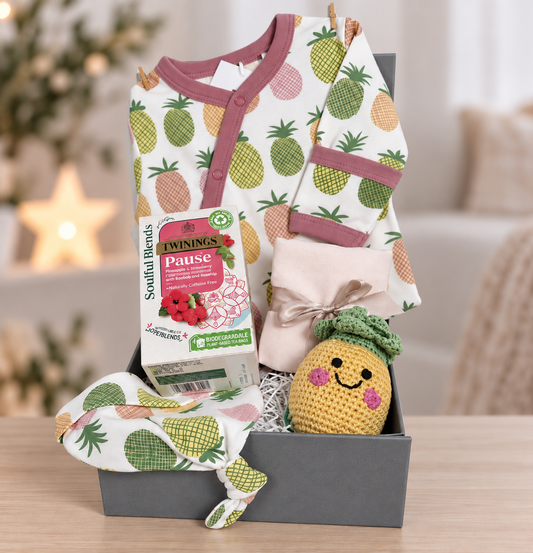 New baby girl hamper containing a organic cotton baby romper with pink, yellow and green pineapple print, matching baby knot hat, a crocheted baby rattle shaped like a pineapple and a box of Twinings teabags.