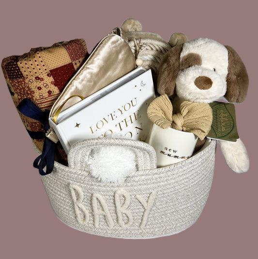 Nappy caddy baby hamper containing a tummy time blanket, baby photo album, ebbs puppy soft toy, new mummy mug, baby headband, baby dressing gown, baby pompom hat in white, a gold coloured silk adult eye mask, a nursery hanging decoration and some baby toiletries.