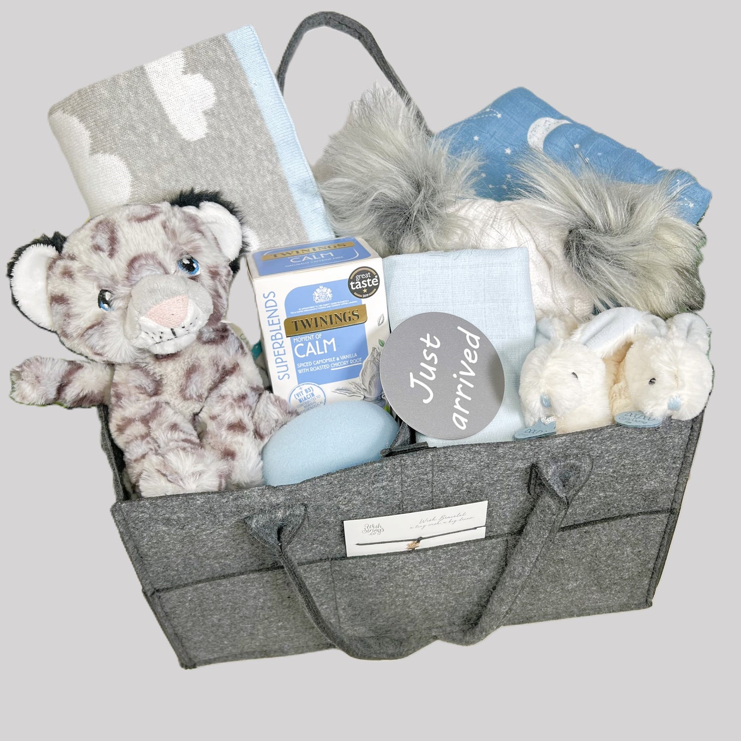A New Baby Boy gift of a dark grey felt nappy caddy containing a cotton baby blanket in grey with a white coloud pattern and sky blue trim, a large blue baby muslin with a white print of planets, a Kelleco snowlepoard soft baby toy, a pair of Doudou Compagnie bunny baby slippers in white and blue, a grey and white baby pompom hat with two fluffy grey pompoms, a packet of Twining Calm teabags, a wish string and various plastic free baby toiletries.