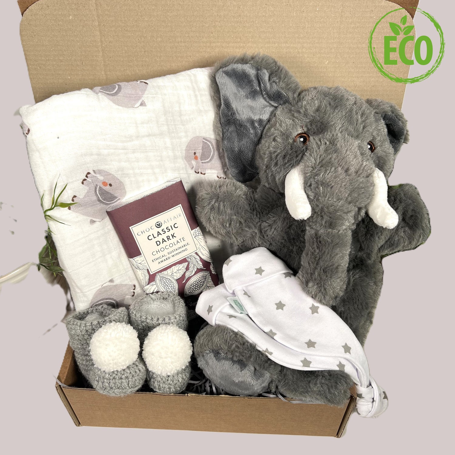 A baby hamper containing a grey elephant puppet made from recycled materials, a white baby knot hat with a grey stars print. A large white muslin square with an elephant print, a pair of crocheted baby bootees in grey with white pom poms and a bar of handmade dark chocolate.