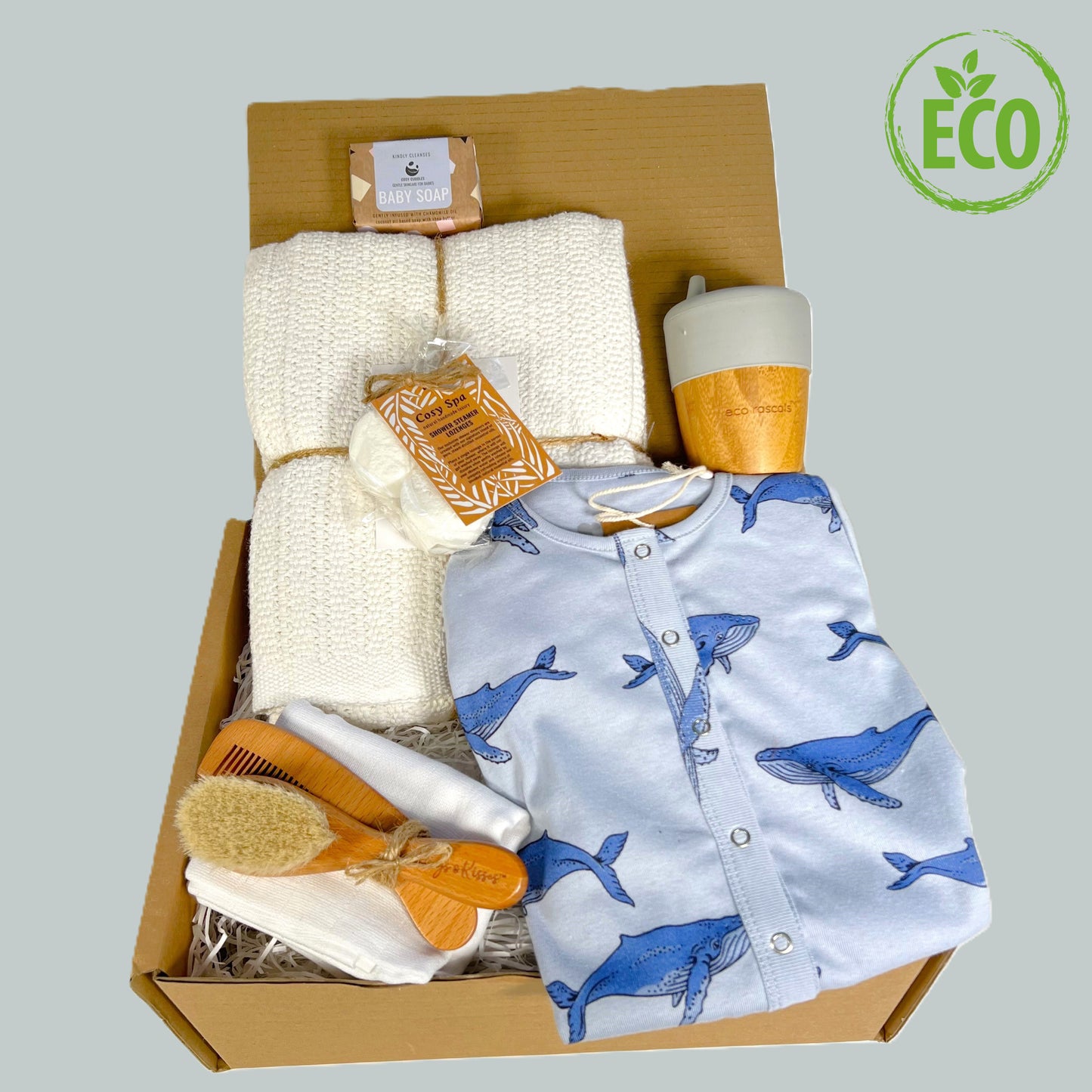 This eco friendly new parents gift has a GOTS certified blue whale print baby romper, a white cotton cellular baby blanket, a bamboo sippy cup by Eco Rascals, a wooden baby brush and comb set, a white muslin square and a pack of spa steamers with refined essential oils.