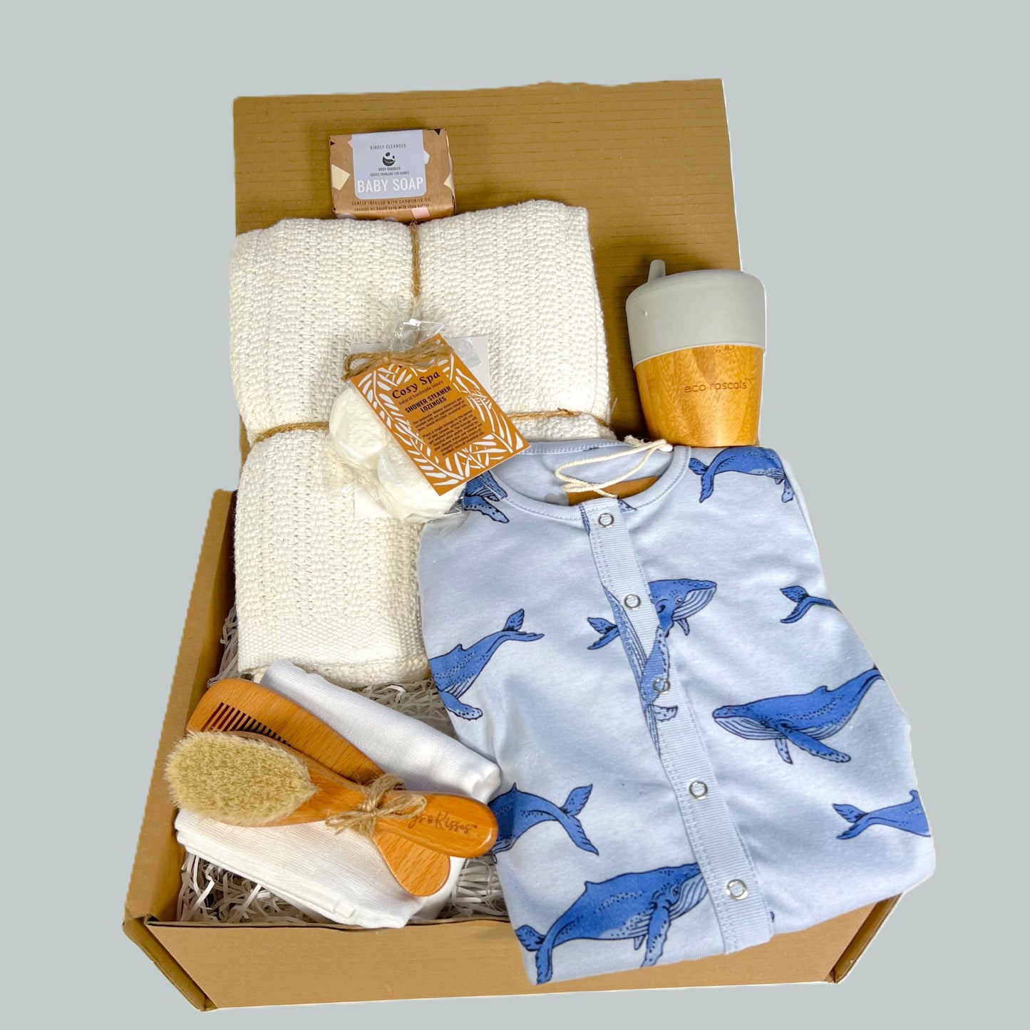 This eco friendly new parents gift has a GOTS certified blue whale print baby romper, a white  cotton cellular baby blanket, a bamboo sippy cup by Eco Rascals, a wooden baby brush and comb set, a white muslin square and a pack of spa steamers with refined essential oils.