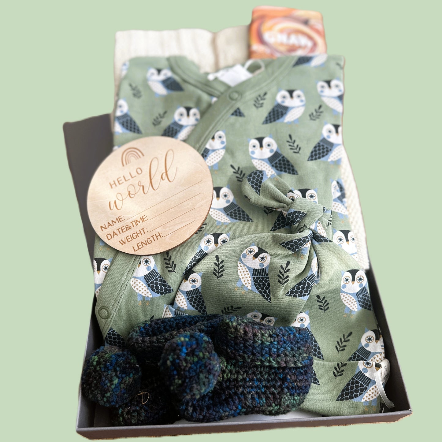 baby hamper with sleepsuit and hat outfit with owl pattern, booties, and 'Hello World' sign in a box on a light green background
