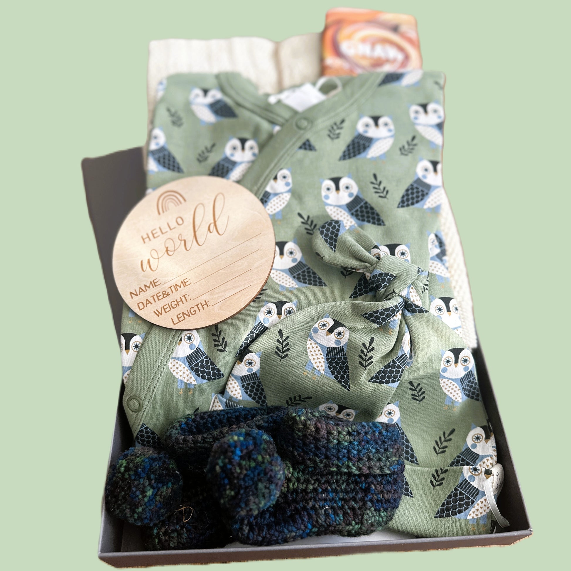 baby hamper with sleepsuit and hat outfit with owl pattern, booties, and 'Hello World' sign in a box on a light green background