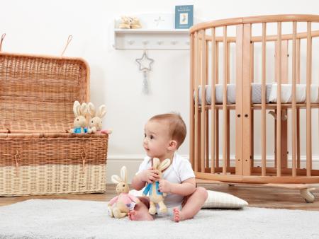 Baby playing with Flopsey bunny baby rattle in a nursery room with a crib and wicker basket.