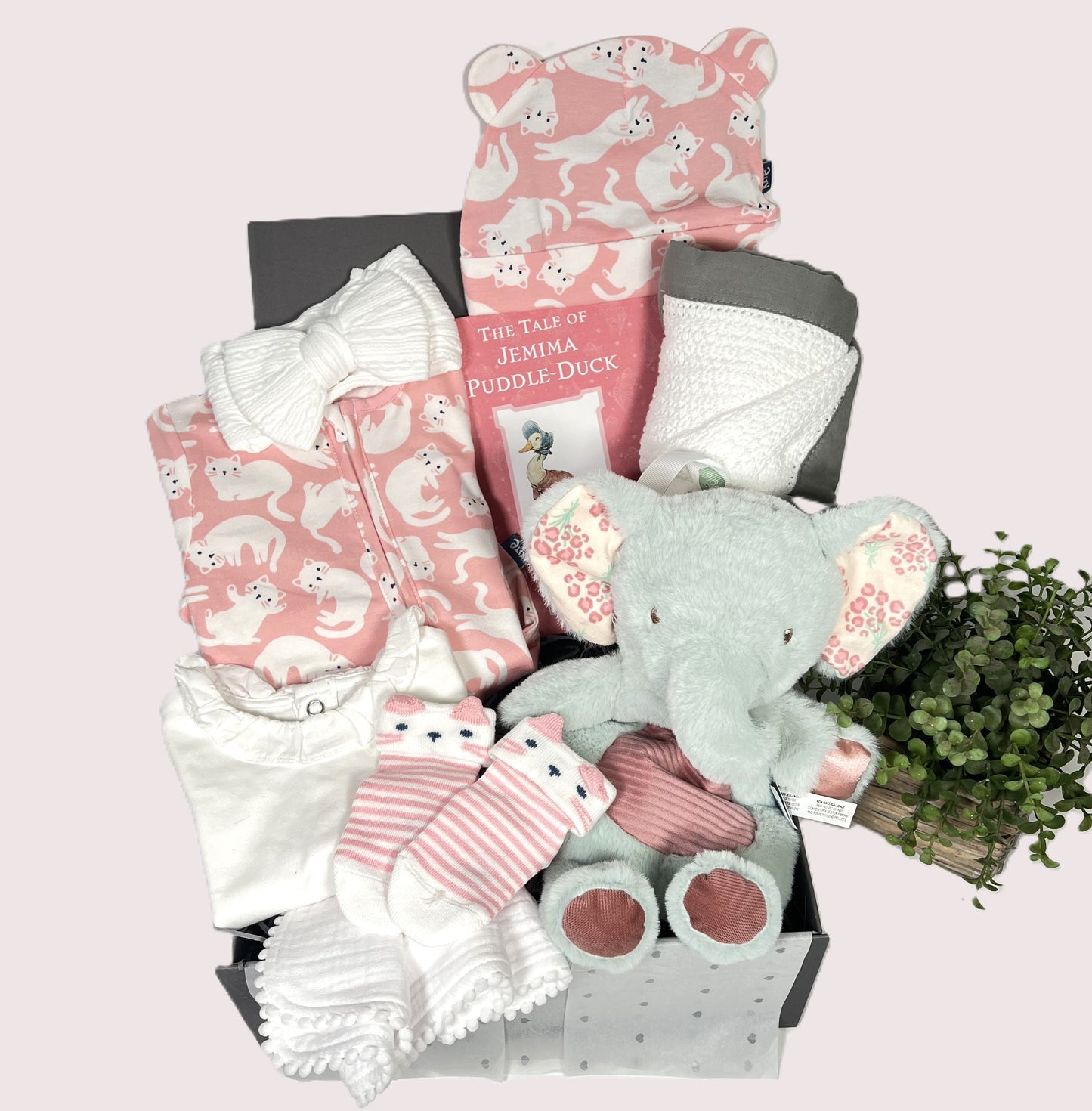 A baby shower gift in a baby keepsake box containing an organic baby sleepsuit in a pink and white kitty print with matching baby hat. A Mary Meyer elephant Lovey soft toy, a white cotton baby blanket and a Jemima Puddleduck reading book.