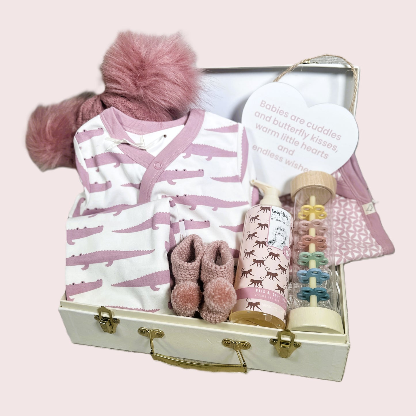 Baby gift set with pink and white outfit, booties, and other items in a suitcase on a light pink background.