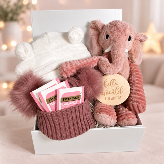 Pink baby girl hamper with a Pink cuddly elephant soft baby toy, a white hooded baby dressing gown, a dusky pink double pompom baby hat, Twinings teabags, a "Hello World" baby photograph sign and a white baby keepsake box.