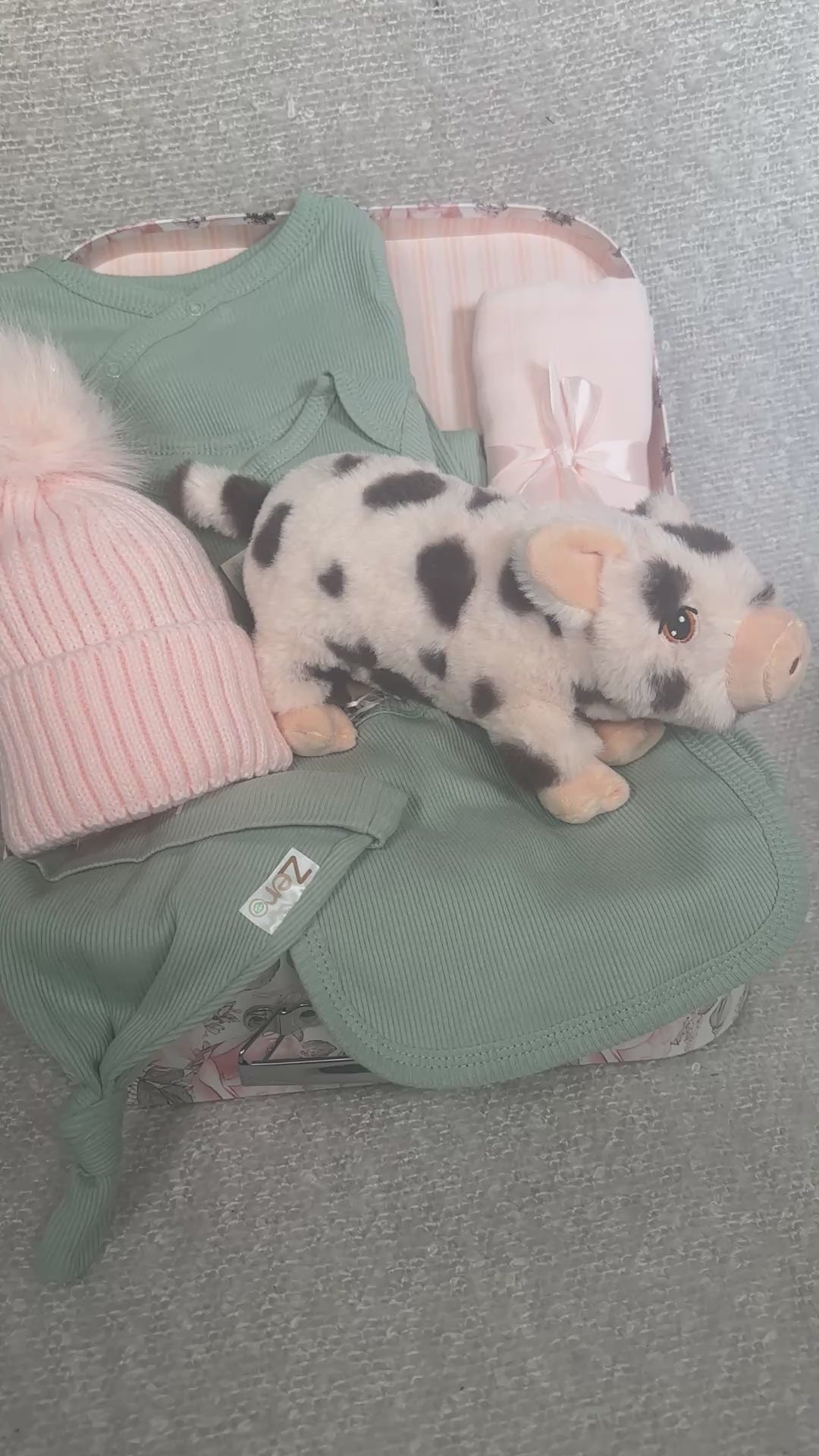 Video of a baby girl hamper with sage green baby clothing, a pink pompom hat and piglet soft baby toy in a keepsake case.