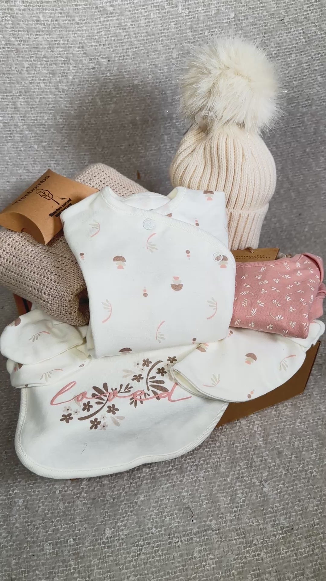 Video of a baby girl hamper with baby clothes, hat, and blanket on a brown background