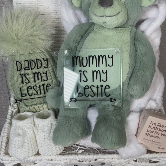 Video of a neutral new baby hamper with a sage green plush monkey toy, a sage green baby pompom hat. Two new parents glass coasters, a pair of baby booties, shower steamer  in a white paper rope hamper.