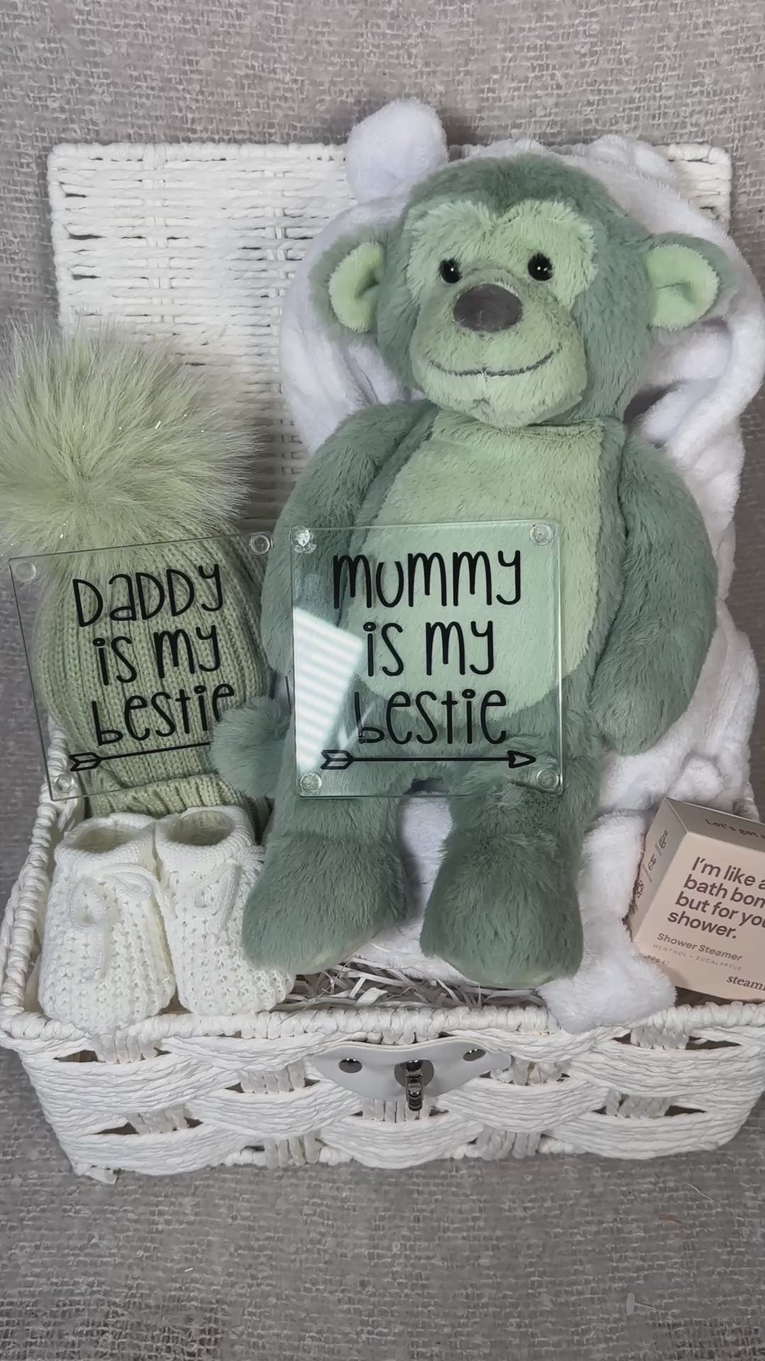 Video of a neutral new baby hamper with a sage green plush monkey toy, a sage green baby pompom hat. Two new parents glass coasters, a pair of baby booties, shower steamer  in a white paper rope hamper.