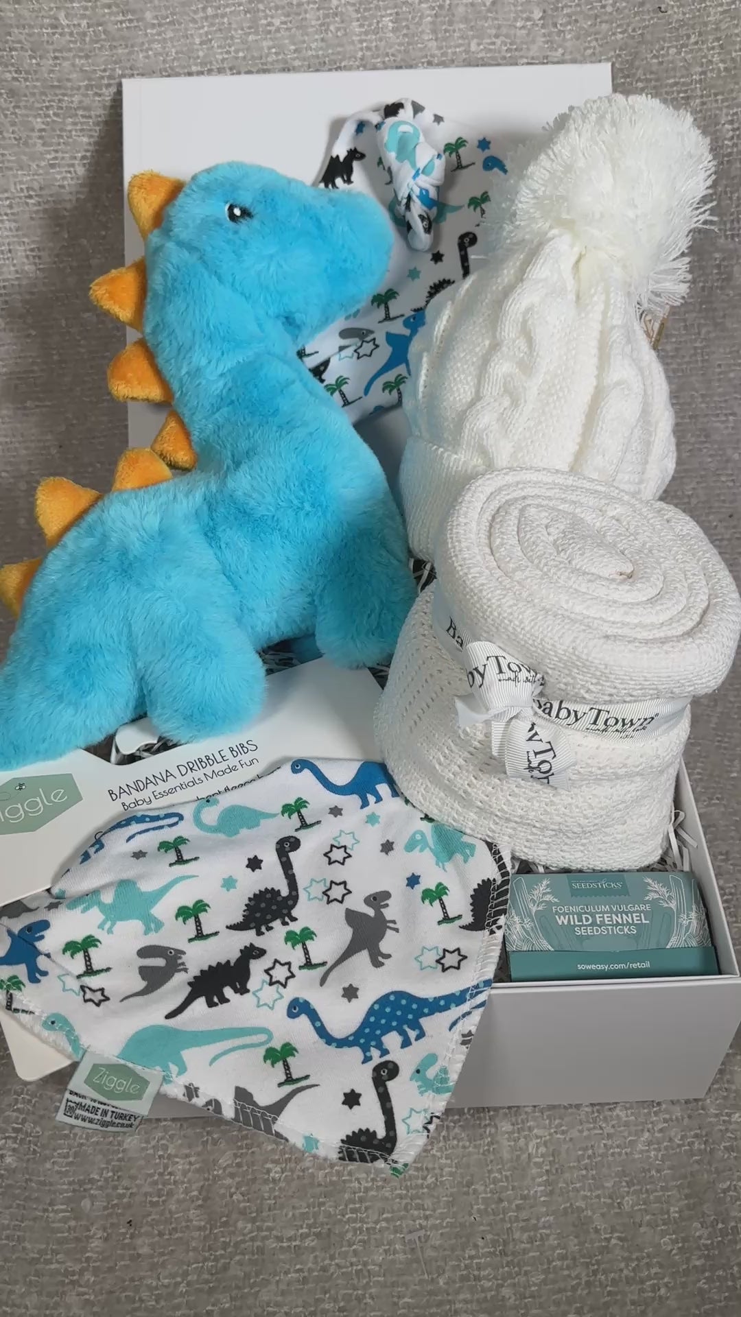 Video of a blue dinosaur new baby hamper with dinosaur toy, baby blanket, dribble bib and baby knotty hat with dinosaur print.