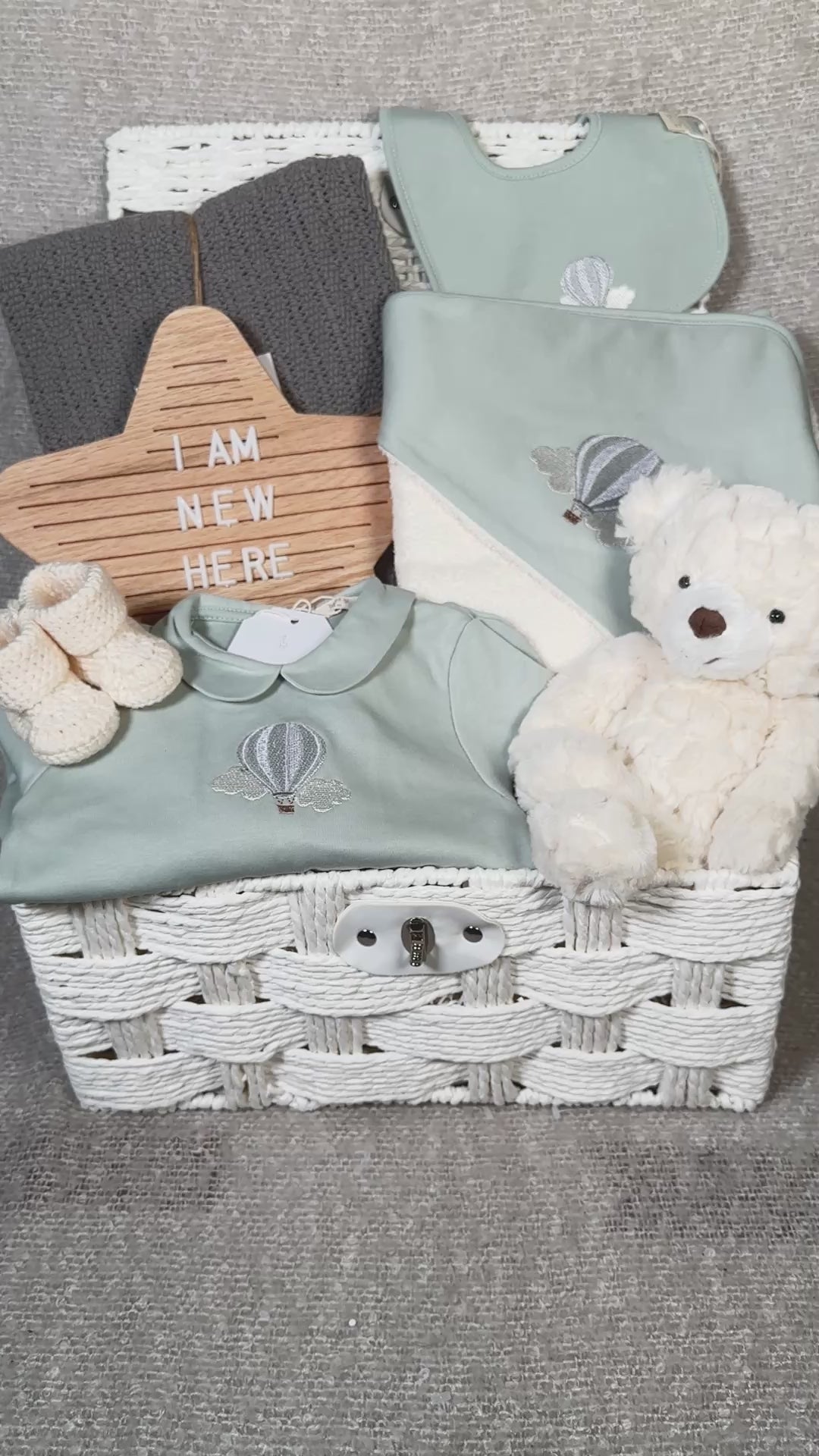 video of a luxury baby hamper in a white paper rope hamper , with sage green baby clothing, hooded baby towel, baby blanket and baby booties
