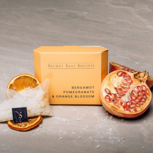 A boxed fragrance product labeled 'Secret Salt Society' and 'Bergamot Pomegranate & Orange Blossom,' styled with a pomegranate half, dried orange slices, and a small salt packet on a gray stone surface.