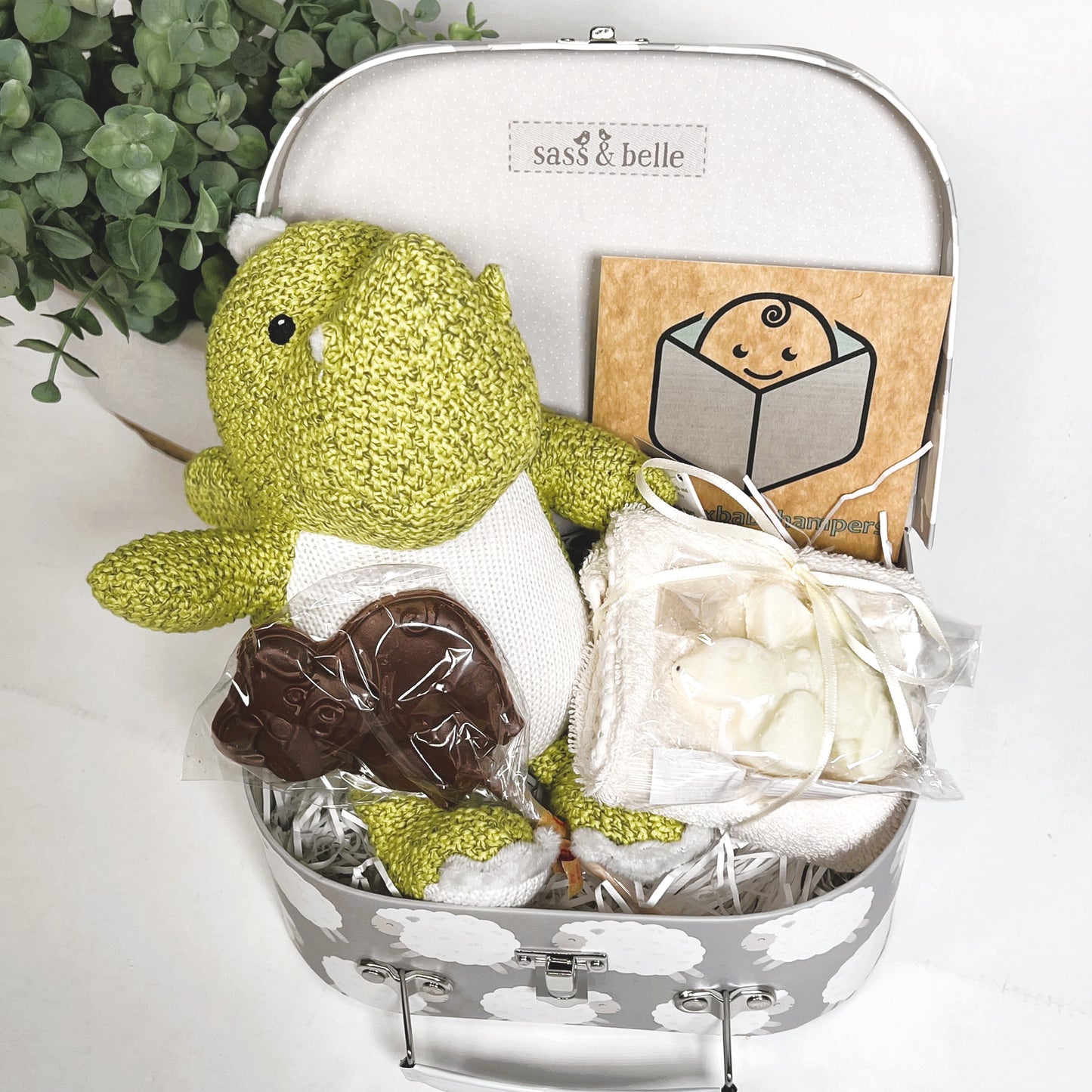 Gift for new siblings in a cardboard suitcase with a sheep print in grey and white containing a lime green dragon baby toy with a white and grey zigzag pattern on it's tummy, a cotton flannel with a bar of stegesorus shaped natural soap and a chocolate lollipop