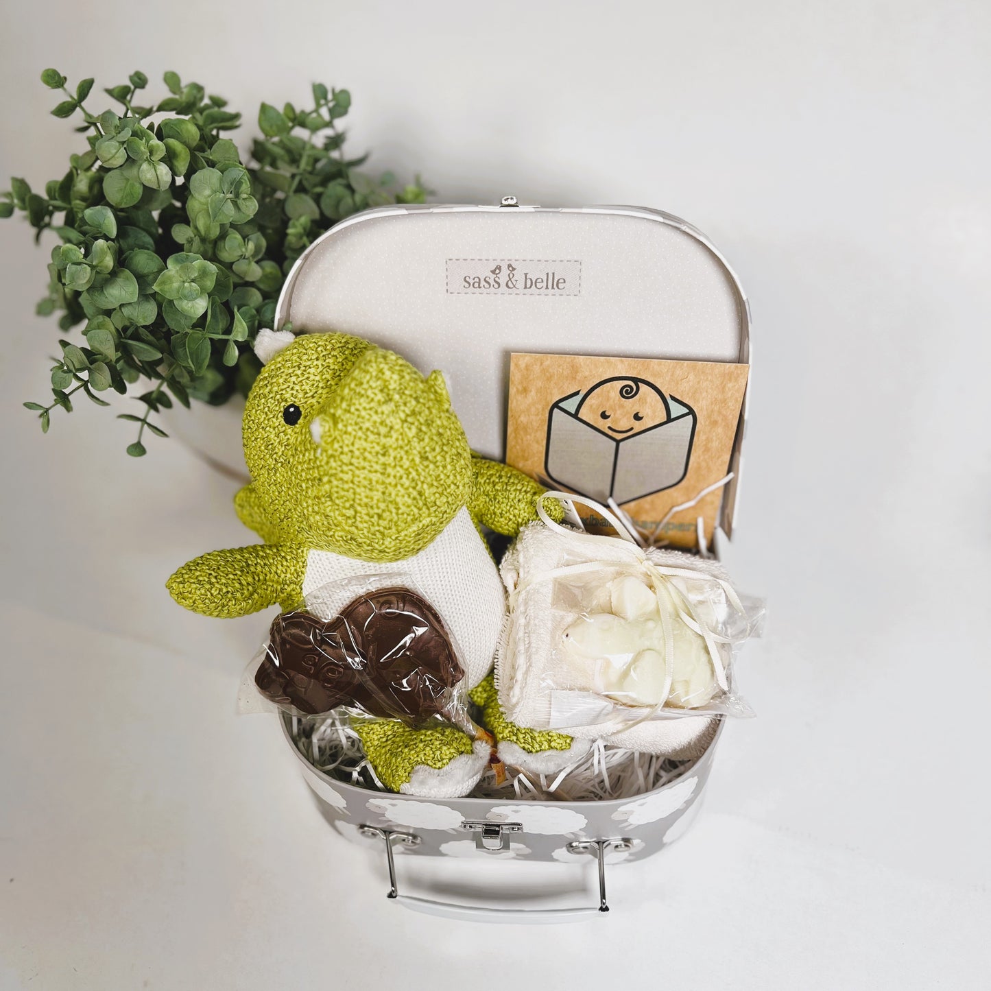 Gift for new siblings in a cardboard suitcase with a sheep print in grey and white containing a lime green dragon baby toy with a white and grey zigzag pattern on it's tummy, a cotton flannel with a bar of stegesorus shaped natural soap and a chocolate lollipop