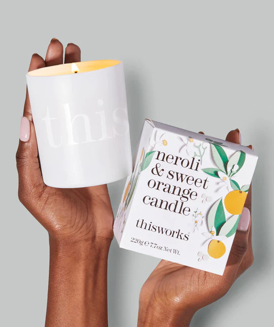 Hand holding a white candle and its packaging labelled 'neroli & sweet orange candle' by 'Thisworks' against a grey background.