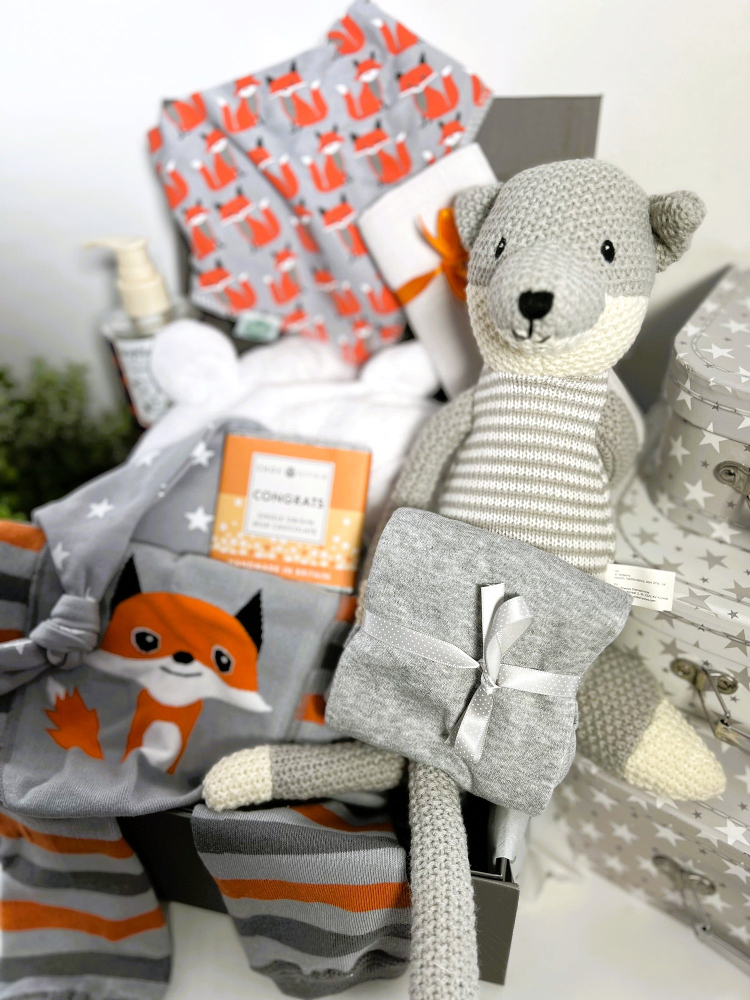 A fox themed new baby hamper containing a wilbeery fox knitted toy, a pair of Ziggle fox baby leggings with a Ziggle fox bib, a white hooded baby dressing gown, a bottle of baby wash, a Ziggle grey and white stars baby knot hat, a muslin square a grey baby vest and a bar of chocolate.