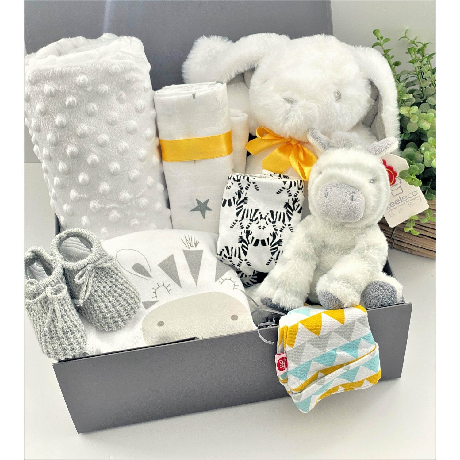 This unisex baby gift hamper comes in a grey magnetic baby keepsake box and contains a large white bunnt baby soft toy with a yellow ribbon around its neck, A zebra soft baby toy, a white baby blanket, a white muslin square with a star pattern, a white baby sleepsuit with a picture od a zebra on the fron that syas "just too cute". A pair of baby booties in grey, a cotton Nuby baby bib with a zebra pattern and a Ziggle geometric pattern baby knot hat.