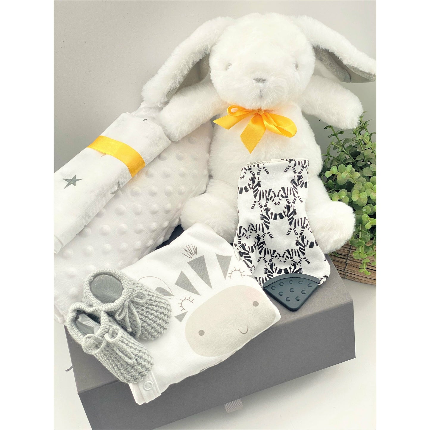 This unisex baby gift hamper comes in a grey magnetic baby keepsake box and contains a large white bunnt baby soft toy with a yellow ribbon around its neck, A zebra soft baby toy, a white baby blanket, a white muslin square with a star pattern, a white baby sleepsuit with a picture od a zebra on the fron that syas "just too cute". A pair of baby booties in grey, a cotton Nuby baby bib with a zebra pattern and a Ziggle geometric pattern baby knot hat.