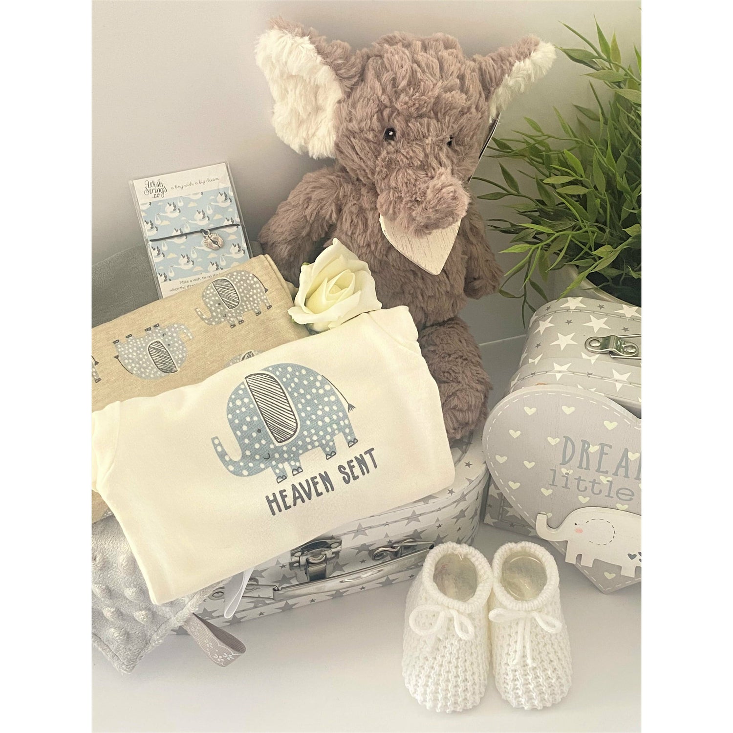 A unisex baby gift hamper in a white and grey stars baby keepsake case containing a Mary Meyer Nursery Putty elephant soft baby toy, a set of 3 coton baby bodysuits with elephant prints and one which reads "Heaven sent", a pair of white baby bootees, a wish braclet for Mum, a heray shaped nursery plaque in grey and white which reads "Dream big little one and has a wooden elephant motif and a grey Taggie blanket.