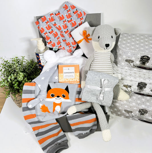 A fox themed new baby hamper containing a wilbeery fox knitted toy, a pair of Ziggle fox baby leggings with a Ziggle fox bib, a white hooded baby dressing gown, a bottle of baby wash, a Ziggle grey and white stars baby knot hat, a muslin square a grey baby vest and a bar of chocolate.