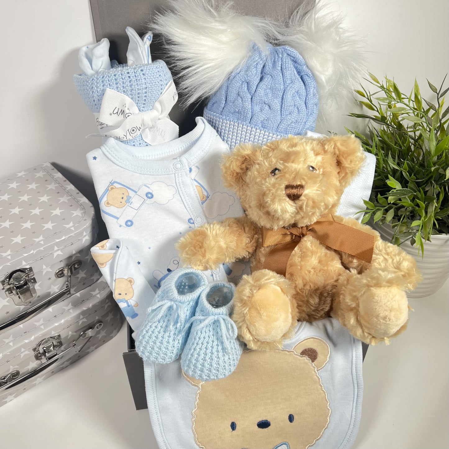 A baby hamper for a boy in a square grey magnetic baby keepsake box containing a blue bear print cooton layette set including a romper suit, baby hat, bodysuit and scratch mitts. A doublpe pompom blue baby hat, a white muslin square a blue cotton cellular baby blanket and a Keeleco Dougie bear with a pair of blue baby bootees.