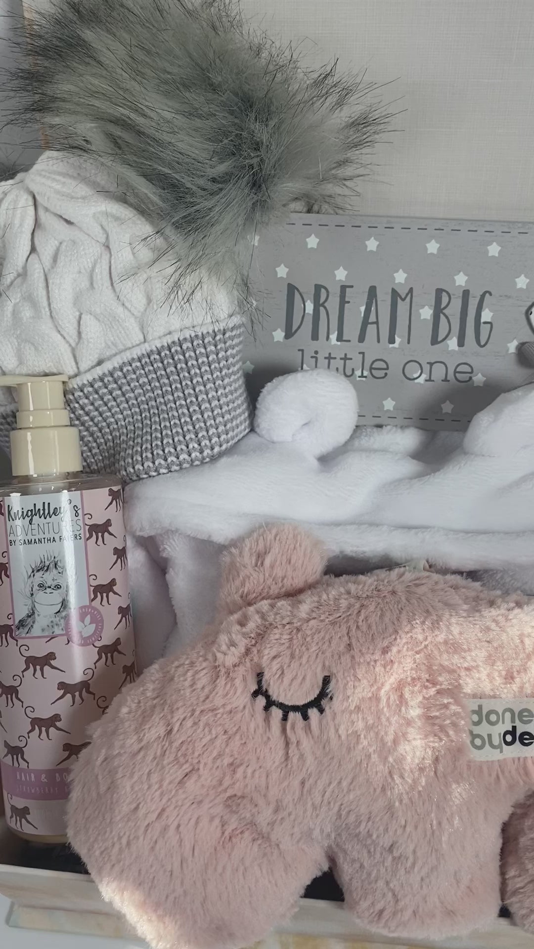 A new baby girl hamper in a magnetic baby keepsake case containing a white baby dressing gown with a hood and bear ears, a white and grey double pom pom baby hat, a pair of white baby booties, a white muslin square, a bottle of baby body wash, a grey and white rectangluar nursery plaque that reads "Dream big little one " and a done By Deer Musical Ozzo baby cuddle in pale pink.