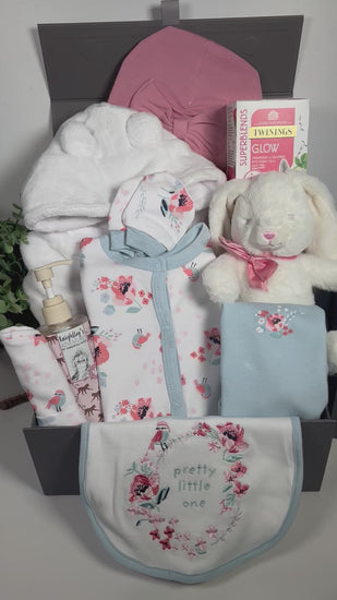 A baby girl hamper in a grey magnetic baby keepsake box containg a 6 piece baby clothing set on white with a blue and pink floral pattern with birds and a blue trim. A matching baby bib, baby vest, hat and scratch mitts.A bottle of baby body wask, a white baby dressing gown, a white bunny baby toy with a pink ribbon, a rose coloured turban baby hat and a pack of Twinings teabags.