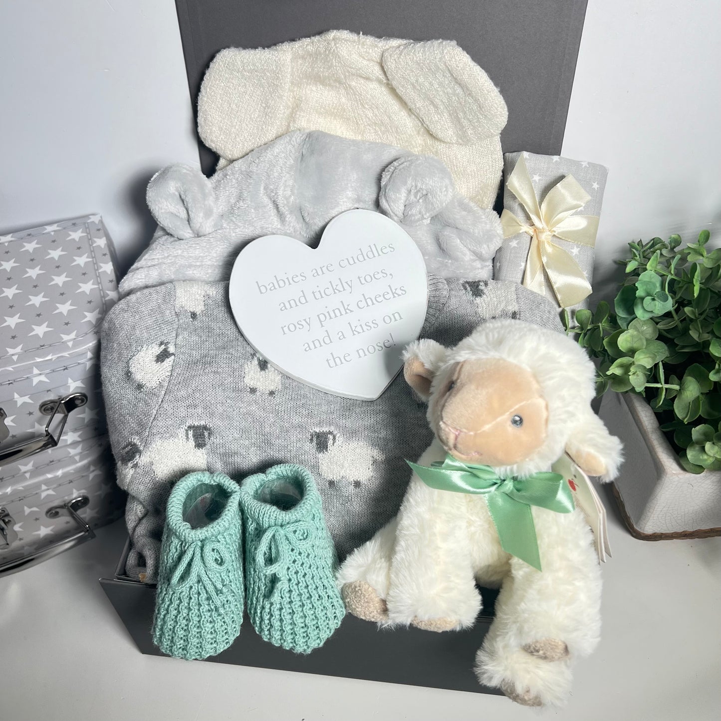 A lamb themed baby hamper in a grey magnetic baby keepsake box with a kniteed gret and white lamb patterned baby romper without feet and a matching lamb eared baby hat. A pair of sage coloured baby bootees, a white heart shaped nursery plaque a cuddly lamb baby toy, a grey and white muslin square a all in a grey magnetic baby keepsake box
