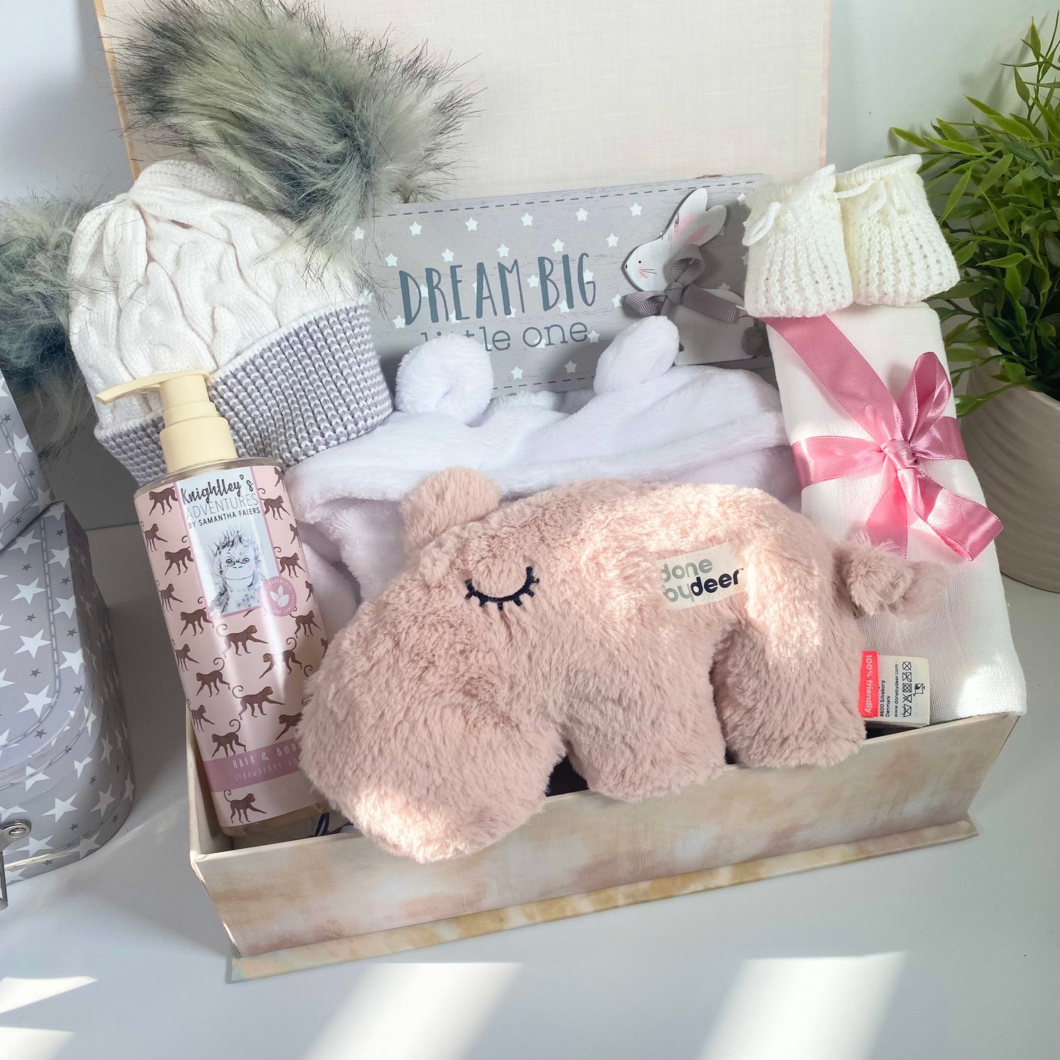 A new baby girl hamper in a magnetic baby keepsake case containing a white baby dressing gown with a hood and bear ears, a white and grey double pom pom baby hat, a pair of white baby booties, a white muslin square, a bottle of baby body wash, a grey and white rectangluar nursery plaque that reads "Dream big little one " and a done By Deer Musical Ozzo baby cuddle in pale pink.
