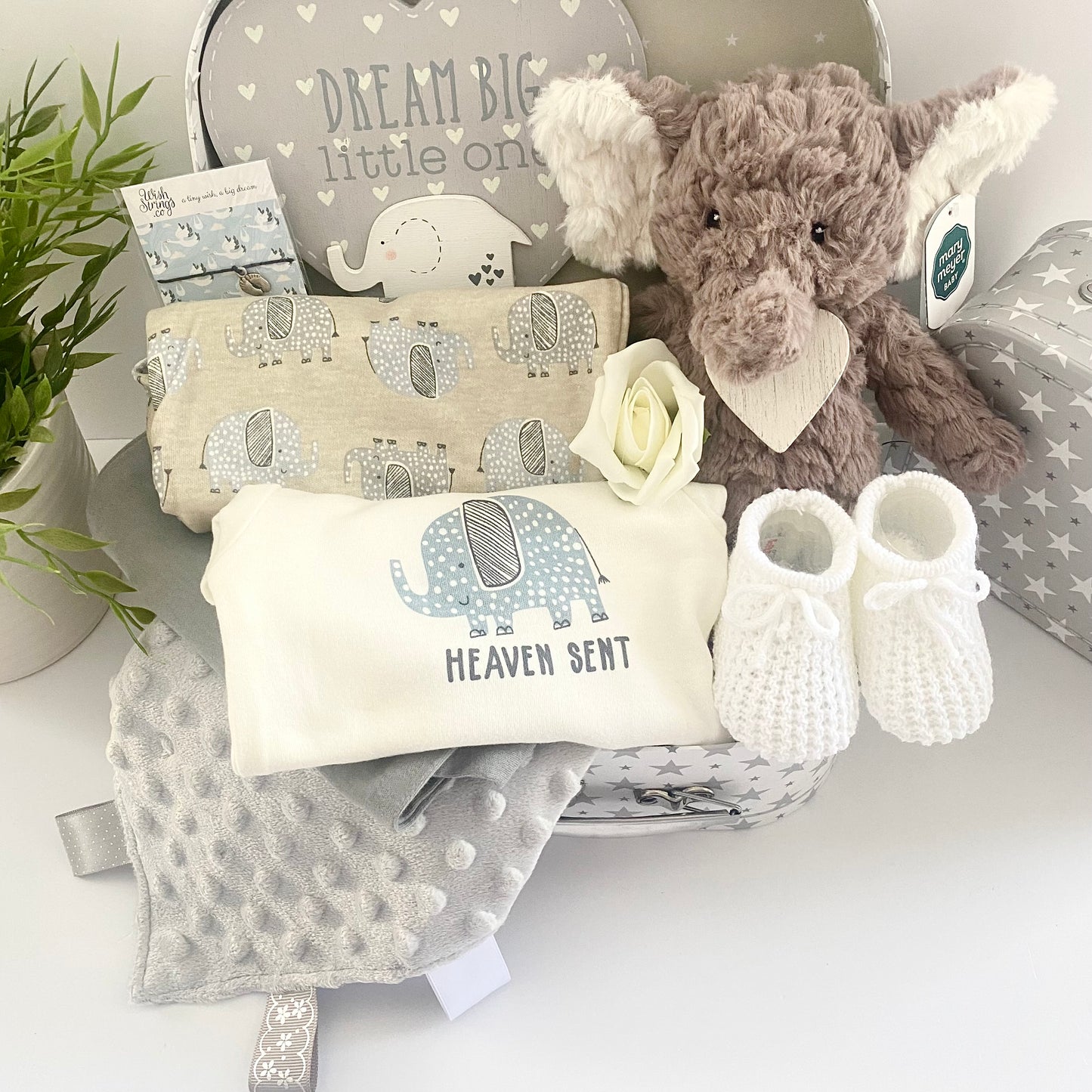 A unisex baby gift hamper in a white and grey stars baby keepsake case containing a Mary Meyer Nursery Putty elephant soft baby toy, a set of 3 coton baby bodysuits with elephant prints and one which reads "Heaven sent", a pair of white baby bootees, a wish braclet for Mum, a heray shaped nursery plaque in grey and white which reads "Dream big little one and has a wooden elephant motif and a grey Taggie blanket.