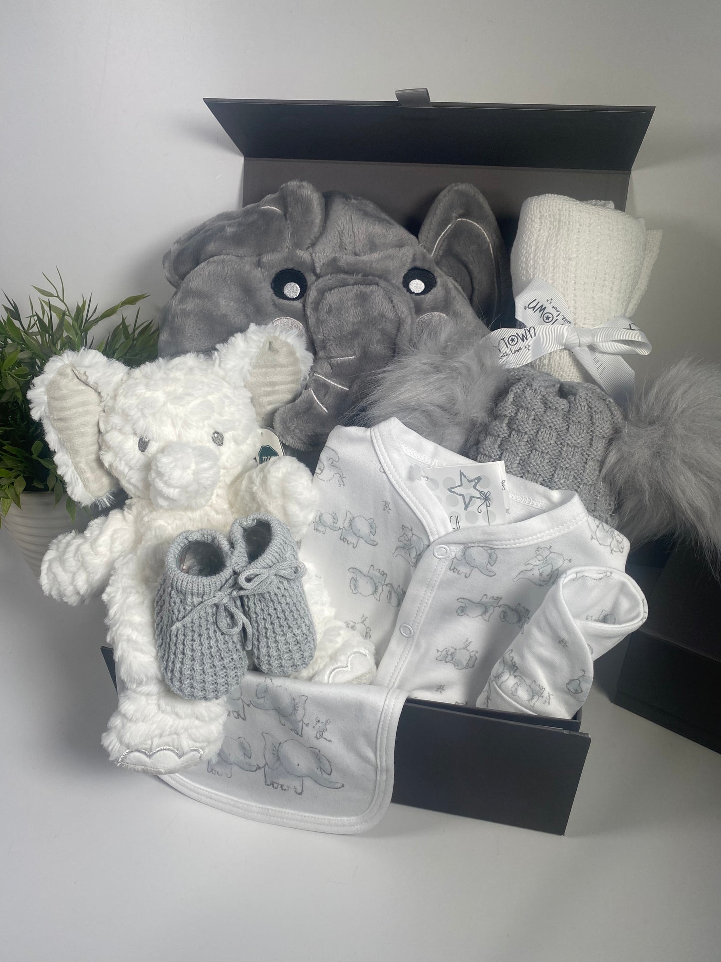 Elephany baby dressing gown, elepahnt baby toy in white, a white sleepsuit with grey elephant print and matching bib and hat,A grey baby dounle pompom hat , a pair pf grey baby booties and a white baby cotton cellular blanket in a baby hamper keepsake box.
