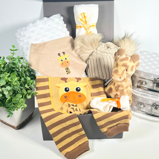 This is a giraffe themed baby gift hamper in a grey magnetic baby keepsake box containing a pair or organge and brown baby leegings with a fun giraffe face on the bottom,  matching cotton baby bid,  biscuit coloured baby pom pom hat, a white soft baby blanket, a white muslin square and a Keeleco 15cm giraffe baby soft toy.