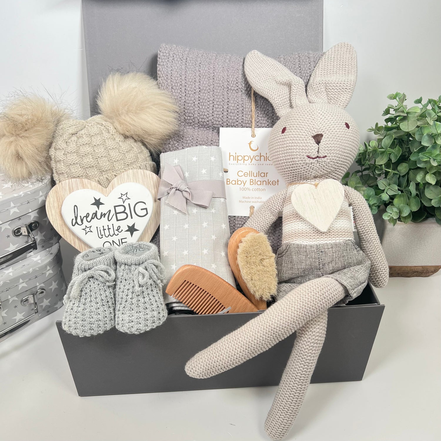 This is a unisex baby gift hamper in a grey magnetic baby keepsake box containg a luxury grey cellular cotton baby blanket, a biscuit coloured baby pom pom hat, a natural wooden baby brush and comb set, a grey muslin square a pair of grey baby bootees, a nursery plaque which reads "Dream big little one" and a grey bunny knitted soft toy.