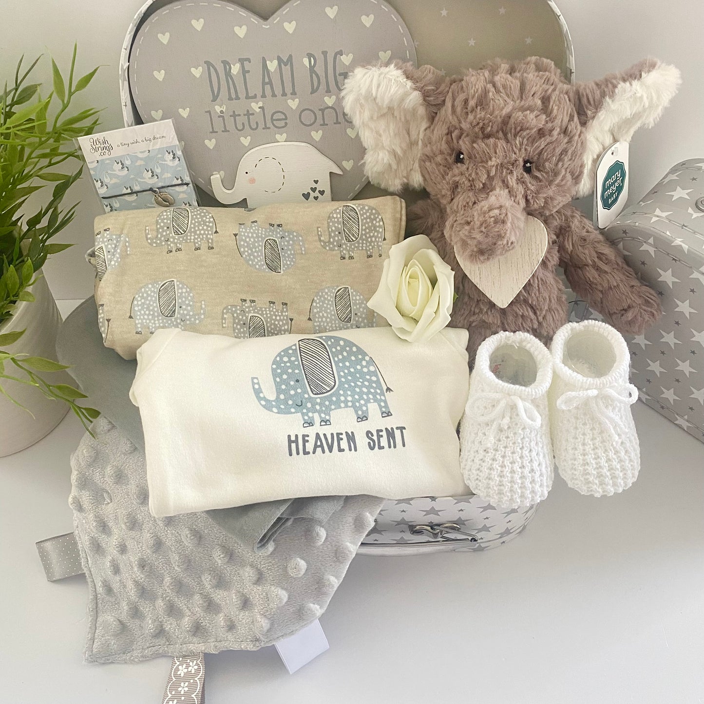 A unisex baby gift hamper in a white and grey stars baby keepsake case containing a Mary Meyer Nursery Putty elephant soft baby toy, a set of 3 coton baby bodysuits with elephant prints and one which reads "Heaven sent", a pair of white baby bootees, a wish braclet for Mum, a heray shaped nursery plaque in grey and white which reads "Dream big little one and has a wooden elephant motif and a grey Taggie blanket.