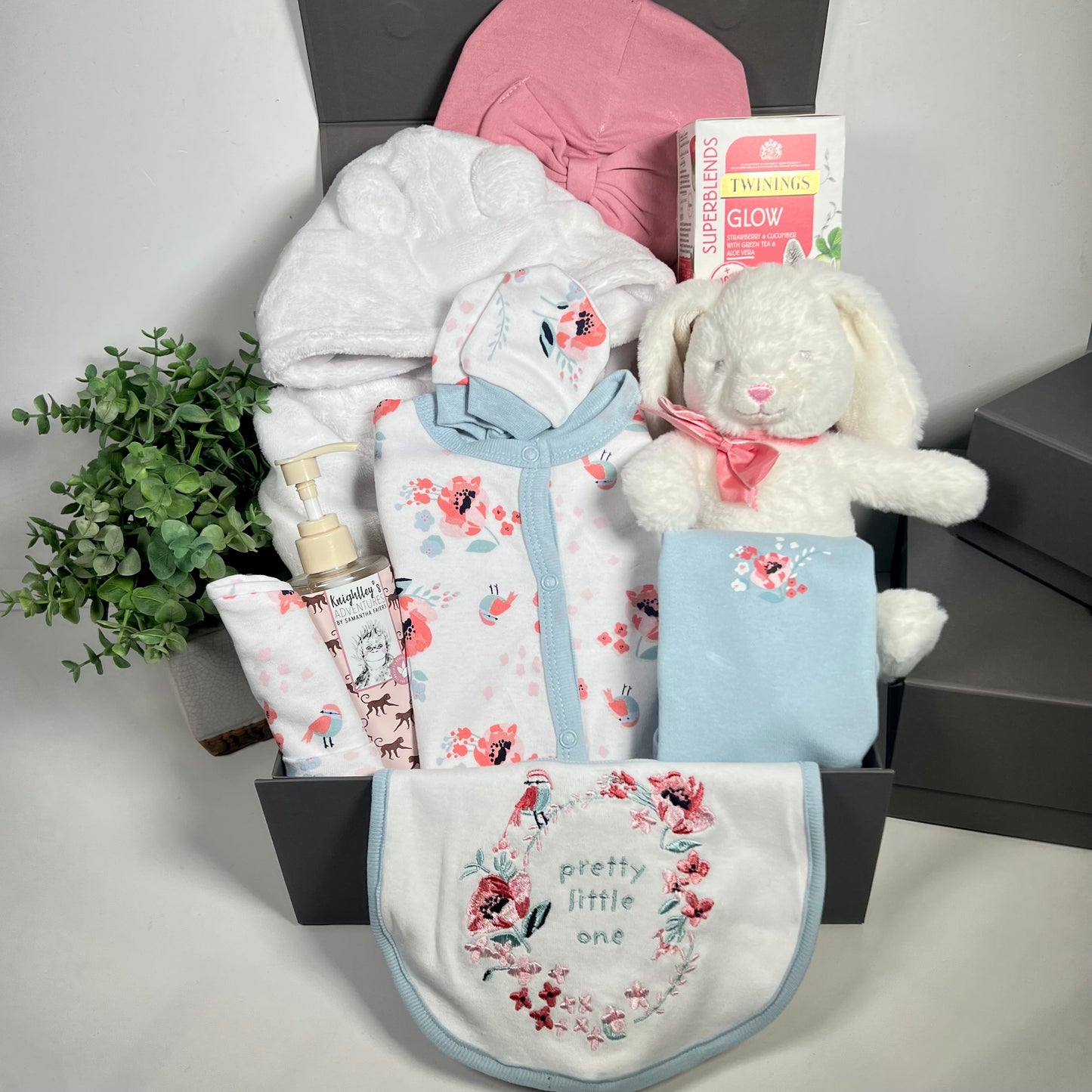 A baby girl hamper in a grey magnetic baby keepsake box containg a 6 piece baby clothing set on white with a blue and pink floral pattern with birds and a blue trim. A matching baby bib, baby vest, hat and scratch mitts.A bottle of baby body wask, a white baby dressing gown, a white bunny baby toy with a pink ribbon, a rose coloured turban baby hat and a pack of Twinings teabags.