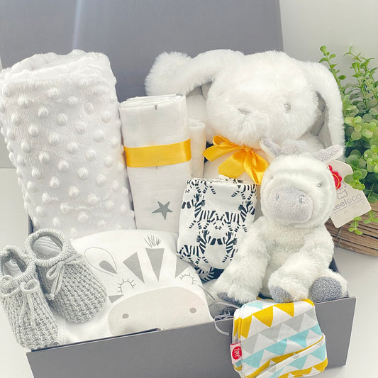 This unisex baby gift hamper comes in a grey magnetic baby keepsake box and contains a large white bunnt baby soft toy with a yellow ribbon around its neck, A zebra soft baby toy, a white baby blanket, a white muslin square with a star pattern, a white baby sleepsuit with a picture of a zebra on the from that reads "just too cute". A pair of baby booties in grey, a cotton Nuby baby bib with a zebra pattern and a Ziggle geometric pattern baby knot hat.