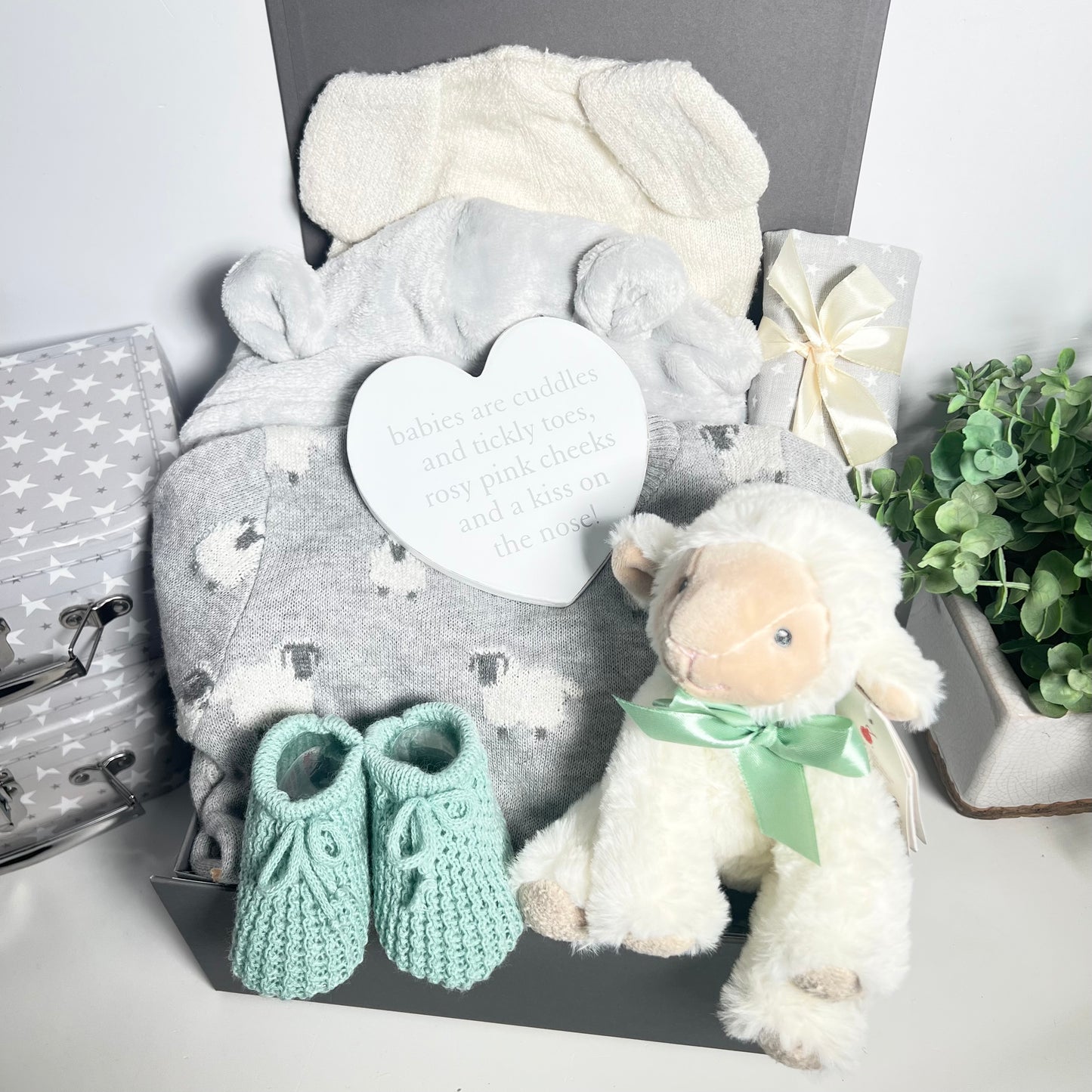 A lamb themed baby hamper in a grey magnetic baby keepsake box with a kniteed gret and white lamb patterned baby romper without feet and a matching lamb eared baby hat. A pair of sage coloured baby bootees, a white heart shaped nursery plaque a cuddly lamb baby toy, a grey and white muslin square a all in a grey magnetic baby keepsake box