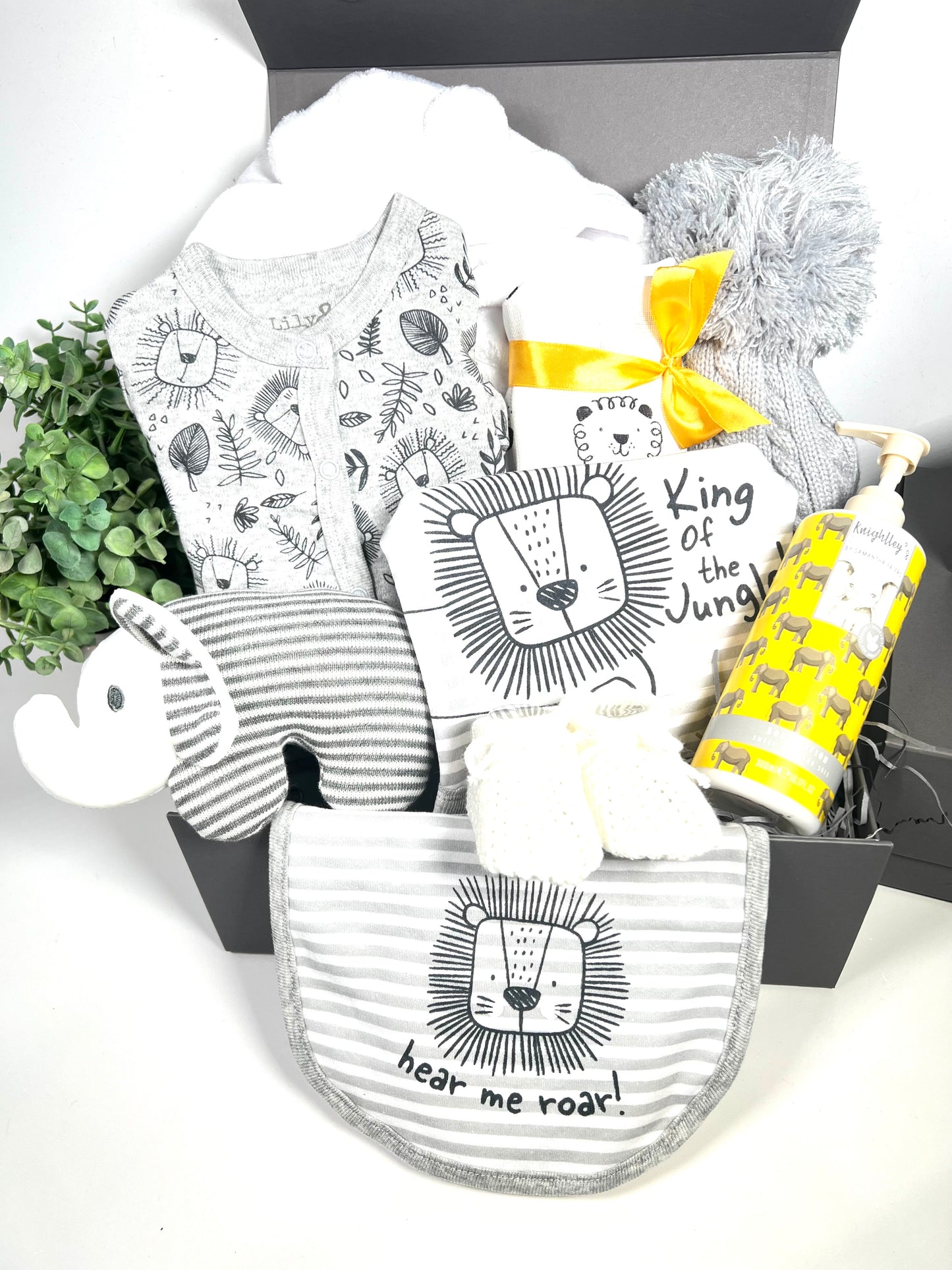 A baby boy gift hamper in a grey magnetic baby keepsake box containing 1 grey baby sleepsuit with a black print of lion faces and jungle leaves in black with a grey and white horizintally striped baby body suit with a picture of a lion and the words "King of the Jungle". There is amatching baby bib withe the words"hear me roar". A botle of vegan baby lotion, a white baby dressing gown, a white baby muslin with lion print, a grey baby pompom hat and a grey and white elephant baby rattle.