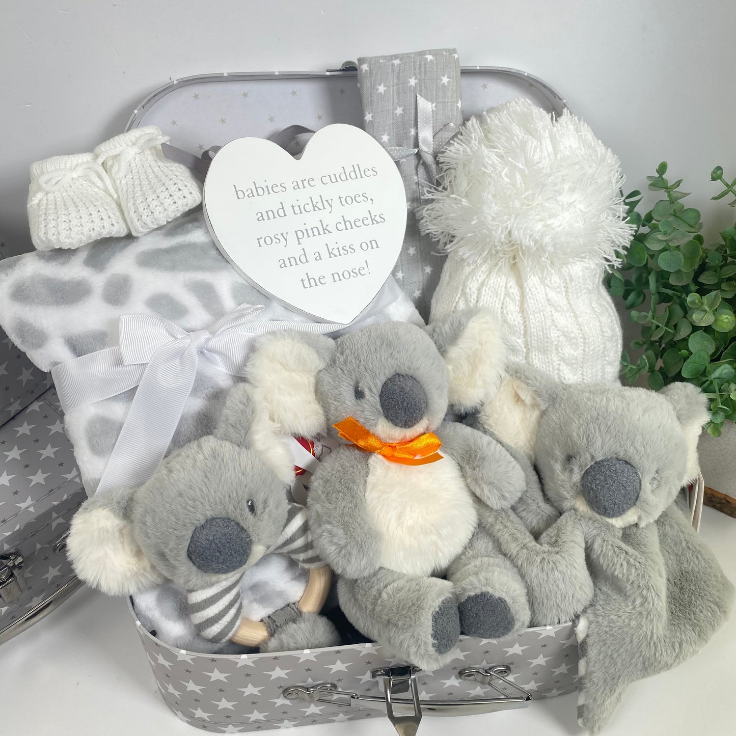 The Koala themed baby hamper comes in a grey and white baby keepsakecase with a pattern of stars. There is a grey and white "giraffe" print baby blanket, white baby pompom hat, a grey and white str pattern muslin square, a pair of white baby booities, a nursery plaque withe the text "babies are cuddles and tickly toes, rosy pink cheeks and a kiss on the nose". There ate three koala bear toys in this baby hamper