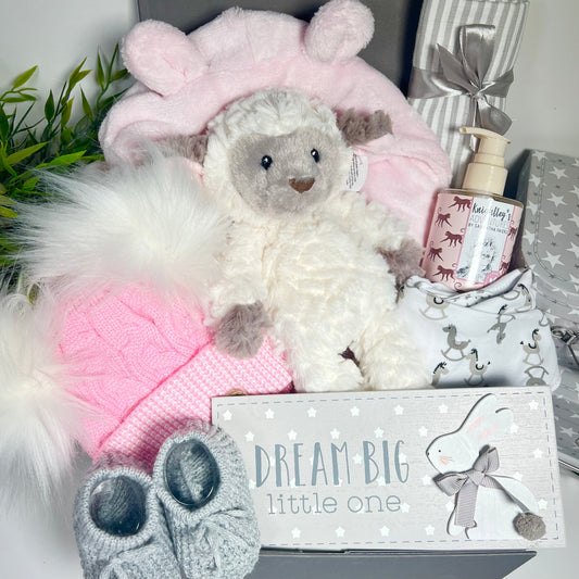 Pink new baby girl gift includes a pink baby dressing gown , a Mary Meyer Lamb soft baby toy, a bottle of baby wash, a grey and white muslin square. apink and white baby pompom hat, a pair of grey baby booties, a baby dribble bib, a nursery plaque all in a grey magnetic baby keepsake case.