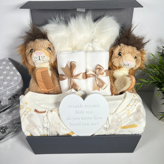 A baby hamper for twins in a grey magnetic baby keepsake box containing 2 x Keeleco lion baby soft toys, 2 x cotton baby sleepsuits, 2 x large white baby muslins, a natural wooden baby brush and comb set, 2 baby pom pom hats and a white heart shaped nursery plaque with grey writing.