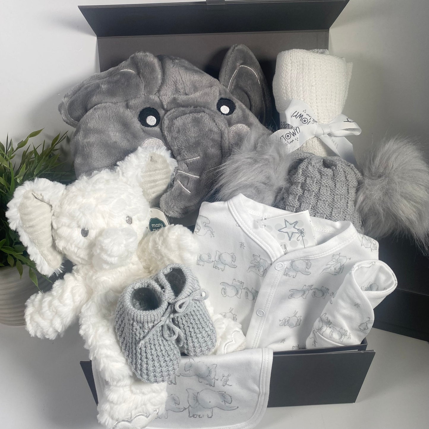 A Grey and white unisex elephant themmed baby hamper in a grey magnetic baby keepsake box with a baby hooded elephant dressing gown, a white Elephant lovey baby soft toy, a sleepsuit with matching bib and hat in white with a grey elephant print. A soft white cotton cellular baby blanakey, a pair of grey baby booties and a baby double pompom hat in grey.