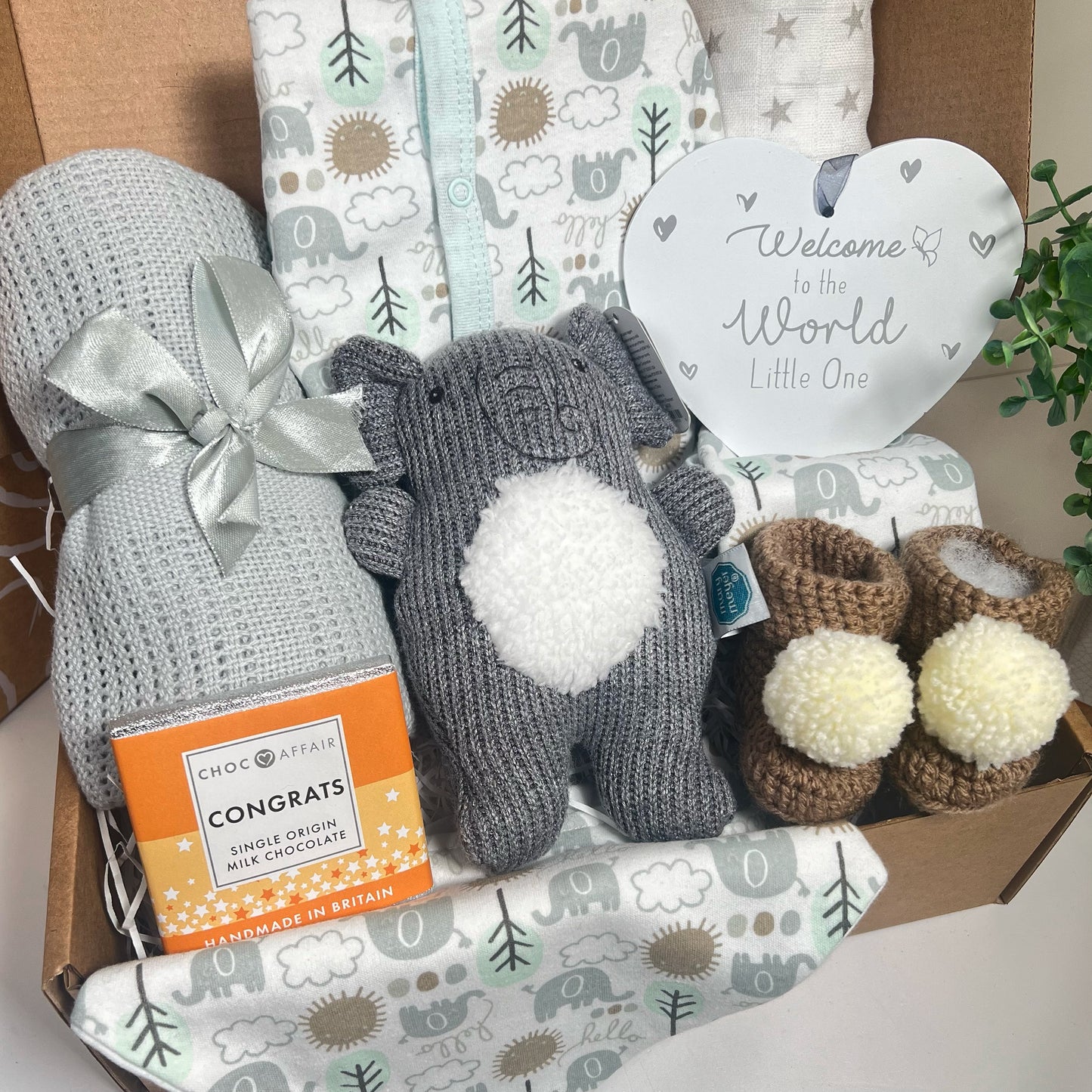 Elephnt themed new mummy hamper contaiing a grey cellular baby blanket , a Mary Meyer elephant baby rattle, a heart shaped nursery plaque, a baby sleepsuit, 2 baby body suits and a matching bib and a bar of chocolate and a pair of chrocheted baby booties with white pompoms.